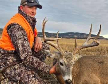 A male SNS Outfitter & Guides client poses in camo clothing and fluorescent orange vest with his 4x6 whitetail buck